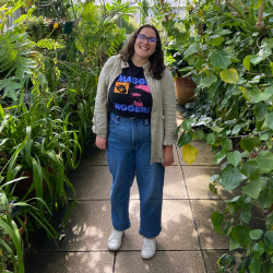 Staff person standing, smiling among green plants. 